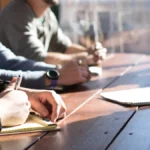 people sitting on chair in front of table while holding pens during daytime